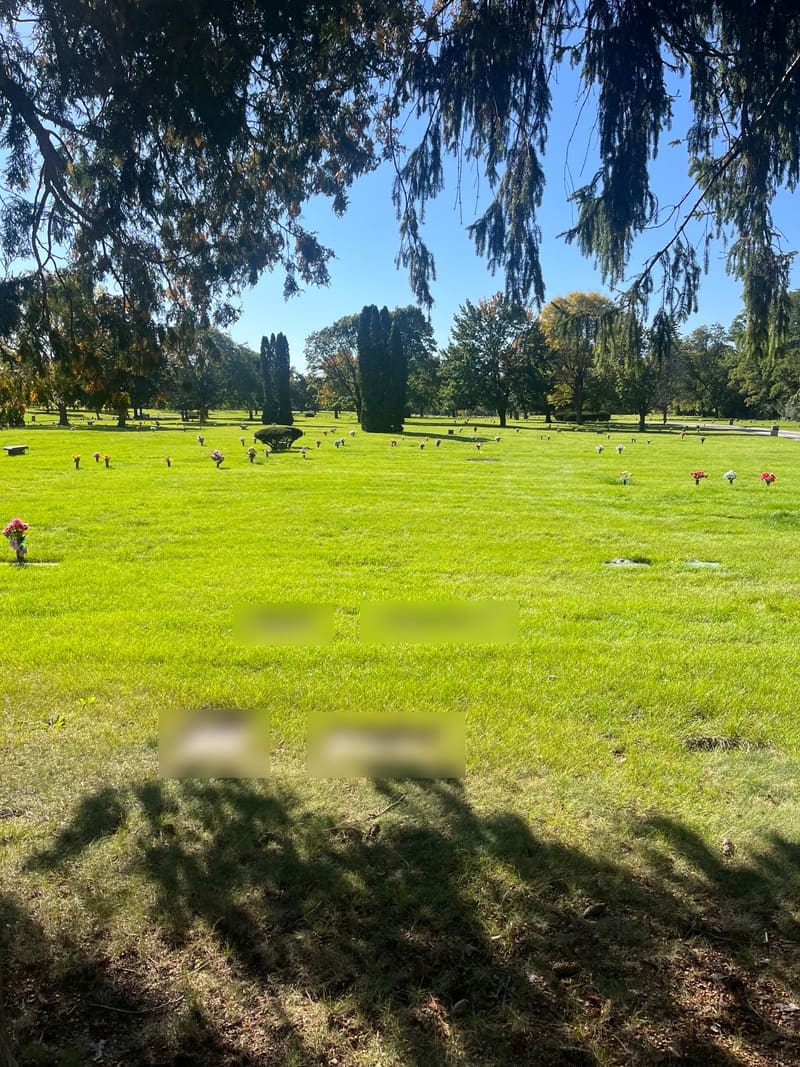 Flat grave markers set into the lawn in the shade of spruce trees. Inscriptions are blurred for privacy.